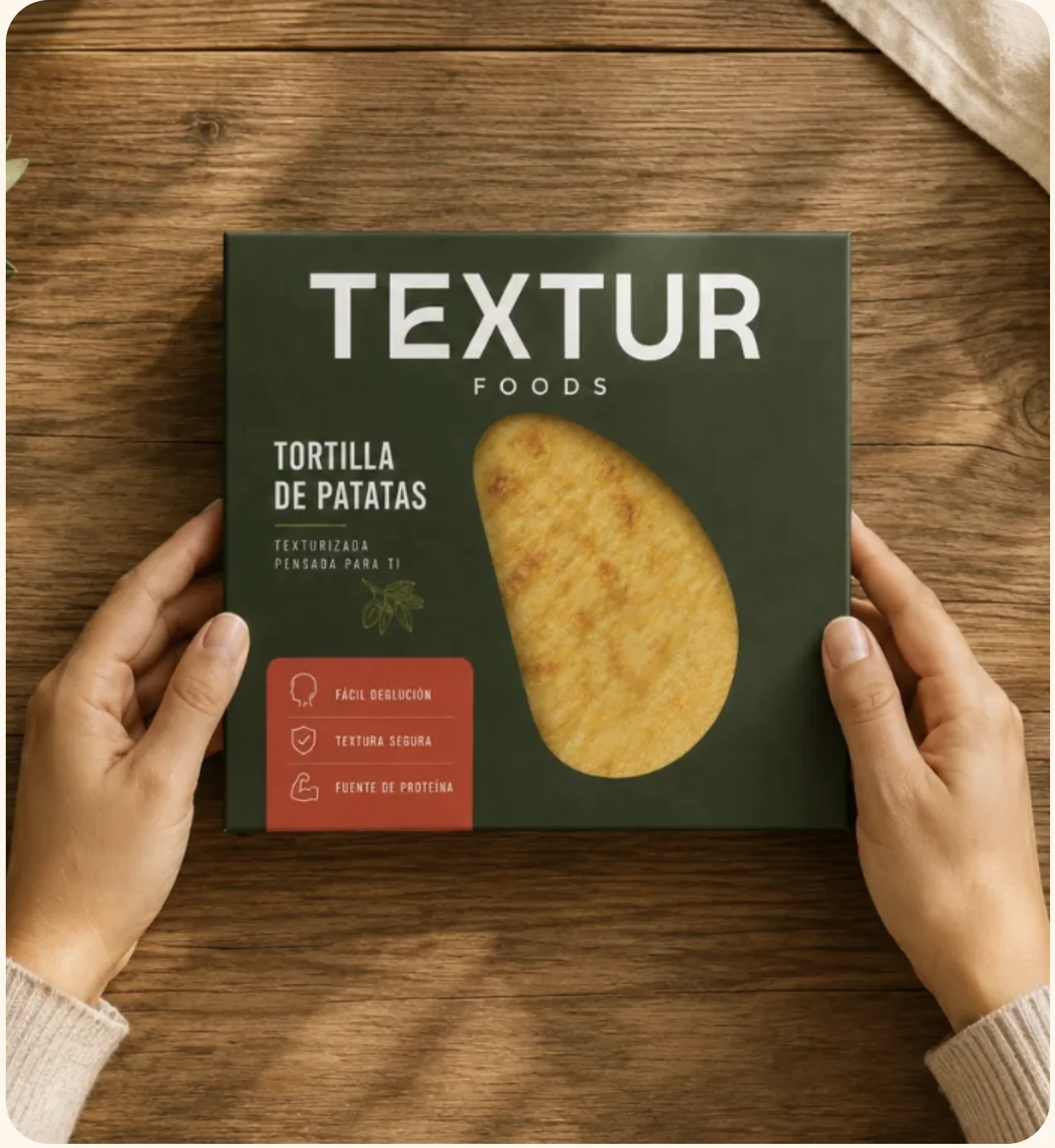 Caja de Textur Foods de tortilla de patatas texturizada sostenida con las dos manos sobre una mesa de madera